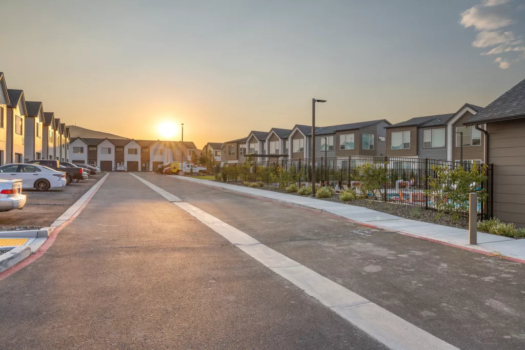 A residential street lined with modern townhouses and parked cars, captured at sunset, with the sun setting in the distance behind the buildings.