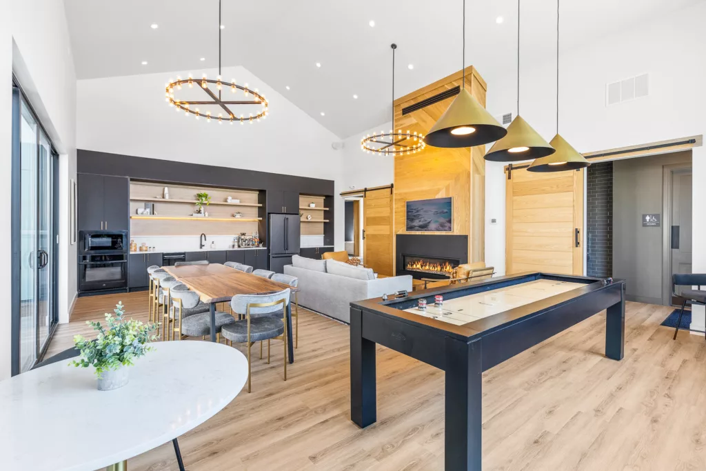 Modern open-plan living room with a large dining table, dark cabinetry, a fireplace, and a shuffleboard table, featuring wooden and black accents with contemporary lighting fixtures.