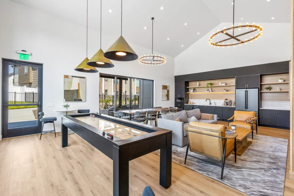 A modern lounge area featuring a shuffleboard table, pendant lights, seating area with sofas and armchairs, a large table with chairs, and a kitchenette with open shelving in the background. The room has high ceilings and a minimalist decor style.