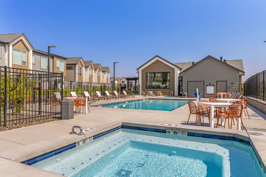 A view of a residential outdoor swimming pool area with sun loungers and tables with chairs under a clear blue sky. The pool is surrounded by a black metal fence, and there are modern-looking homes in the background.
