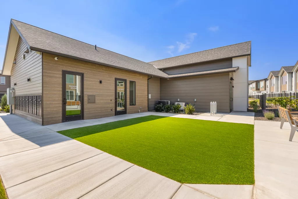 A modern building with a sloped roof and wood paneling, featuring doors labeled 'Package Room' and 'Pet Wash'. The foreground shows a well-maintained lawn and concrete walkway, with additional buildings in the background under a clear blue sky.