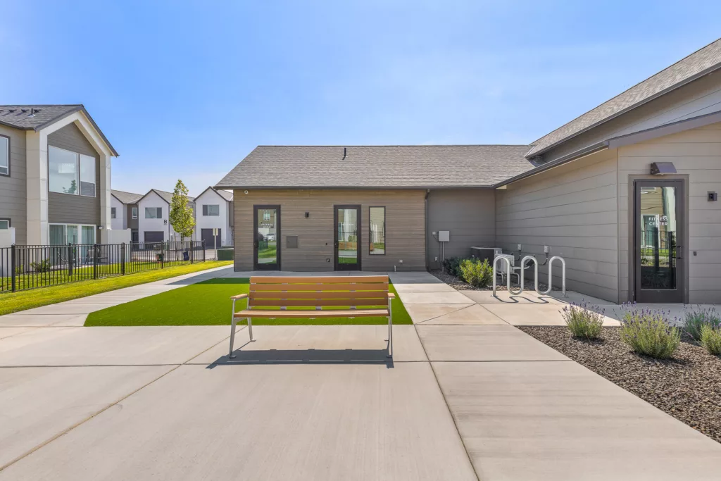 A modern residential complex courtyard featuring a bench, manicured lawns, and a fitness center with glass doors, surrounded by multi-story buildings.