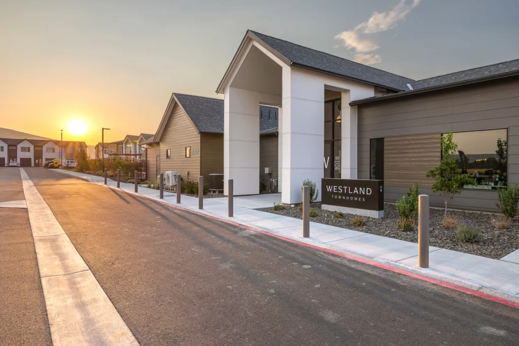 A modern townhouse complex named "Westland Townhomes" at sunset, with a main building featuring a slanted roof and a clear sign in front, surrounded by landscaped pathways and other residential structures in the background.