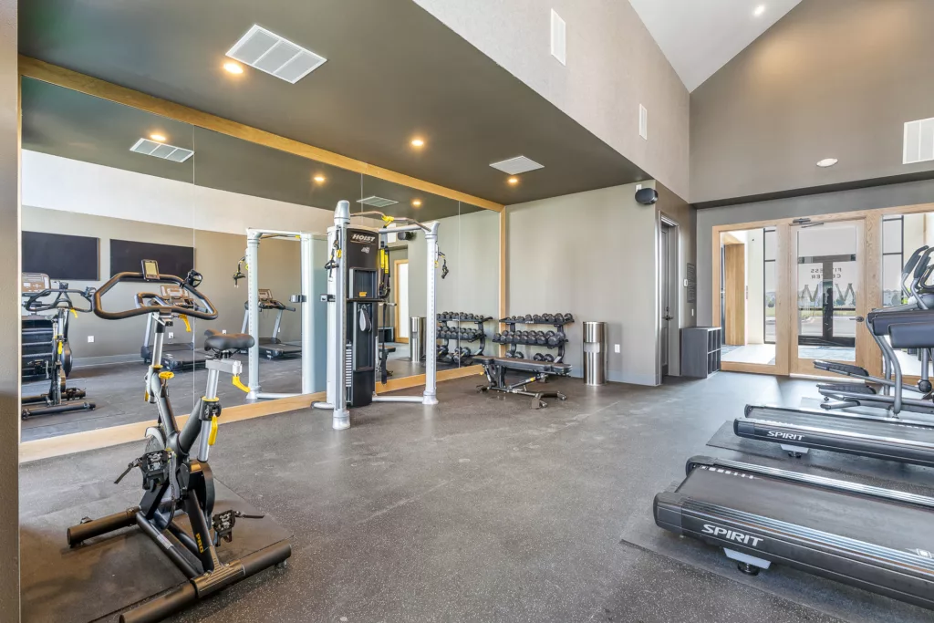 A modern gym interior featuring treadmills, an exercise bike, a multi-function weight machine, and dumbbells on a rack, with a large mirror on the wall and polished floors.