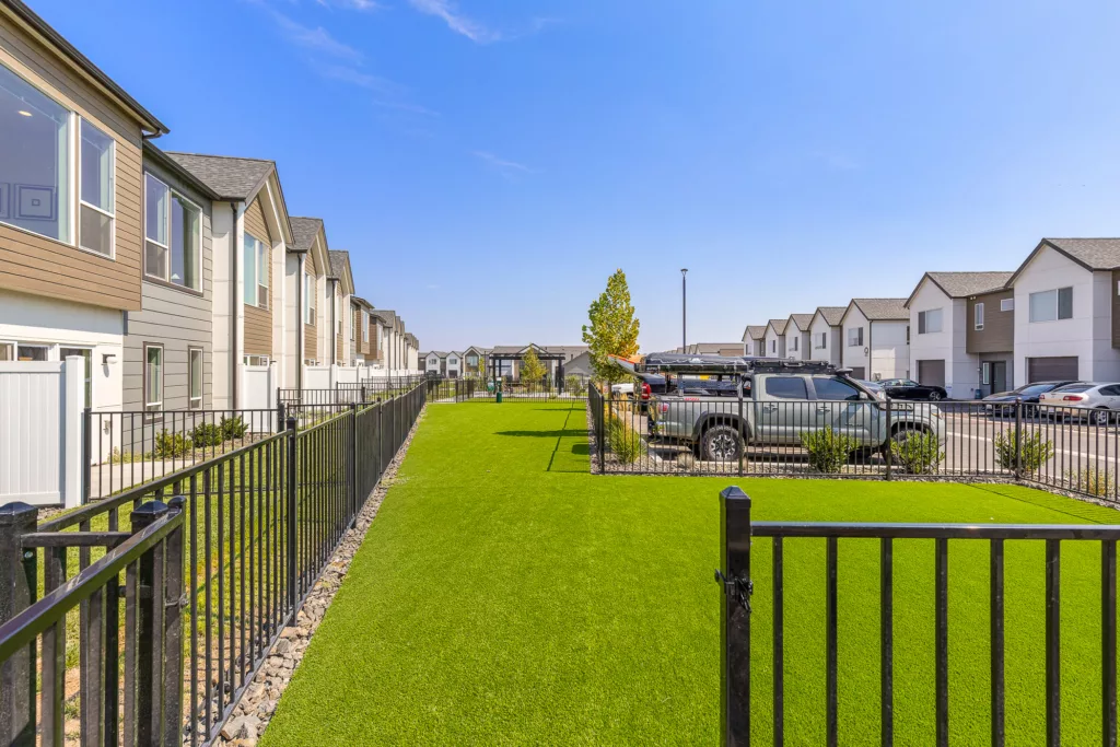 A row of modern townhouses with a black metal fence and a strip of green lawn separates the houses from a parking area on a clear, sunny day.