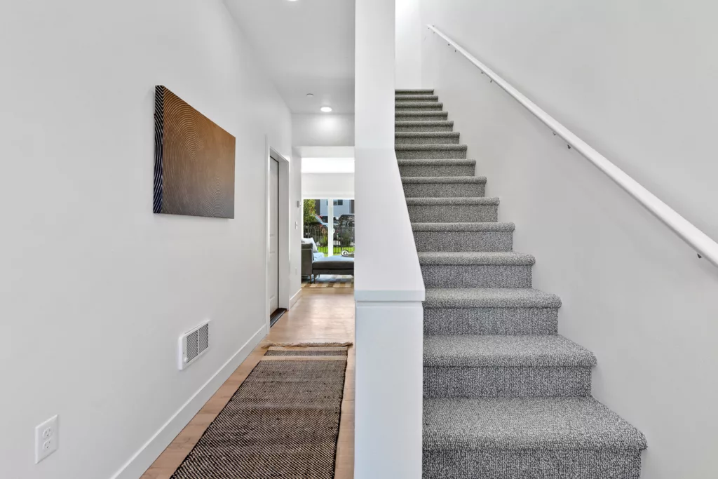 A modern hallway with a gray carpeted staircase on the right and a long rug on the wooden floor extending towards a brightly lit living room in the background.