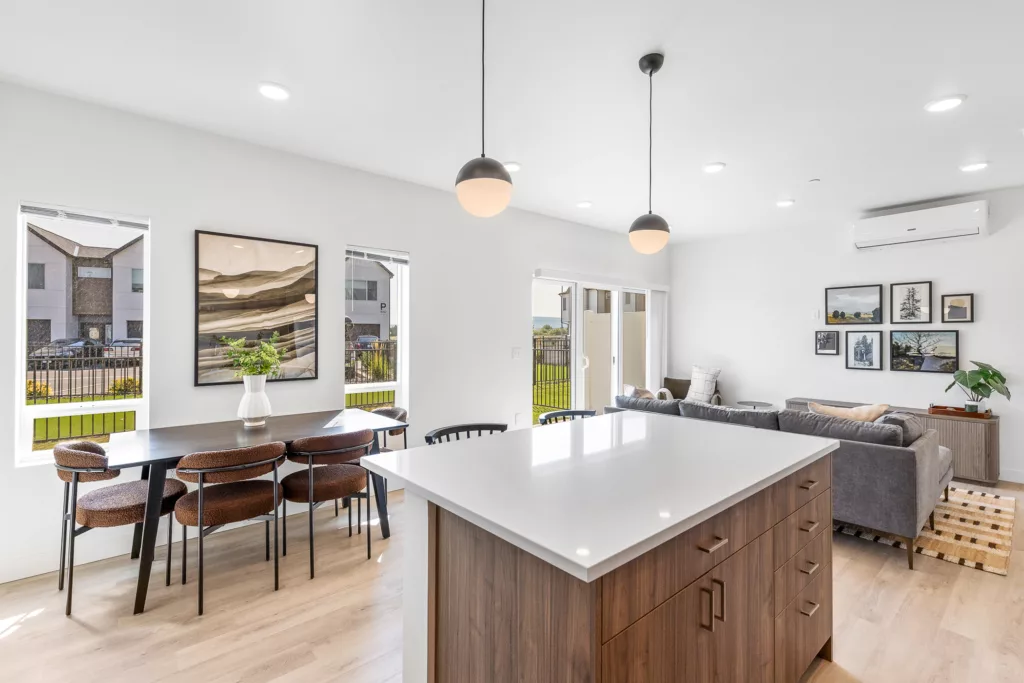 Modern open-concept living and dining area with a kitchen island in the foreground, a dark wooden dining table with chairs to the left, and a gray sectional sofa near the far wall. The room features large windows, pendant lights, and framed artwork on the walls.