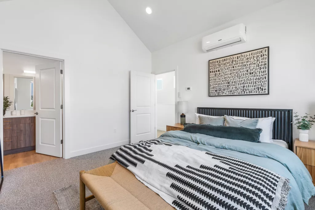 A modern bedroom with a neatly made bed featuring a blue and white color scheme, a patterned throw blanket, and decorative pillows. The room includes a wooden nightstand with a lamp, an air conditioning unit mounted on the wall, abstract artwork, and a view into an adjoining bathroom through an open door.