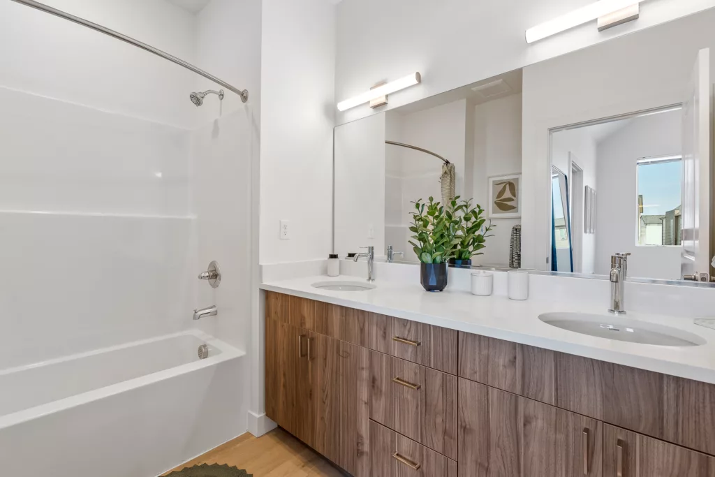 A modern bathroom with a white bathtub-shower combination on the left, a wood-grain vanity with dual sinks and a quartz countertop on the right, and a large mirror above the sink area. A plant in a black pot and two small containers are placed on the countertop, and wooden cabinets with metal handles provide storage.