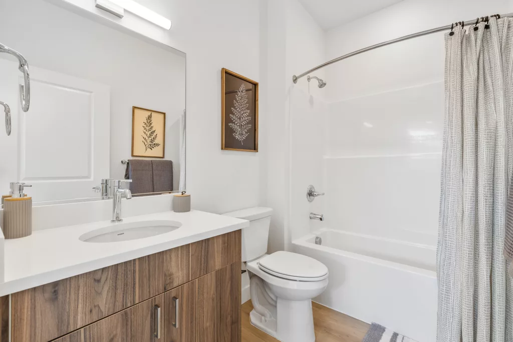A modern bathroom featuring a wooden vanity with a white countertop and sink, a large mirror, a toilet, and a bathtub with a shower curtain. The walls are white, and there's a framed botanical artwork above the toilet. A striped rug is placed on the floor.