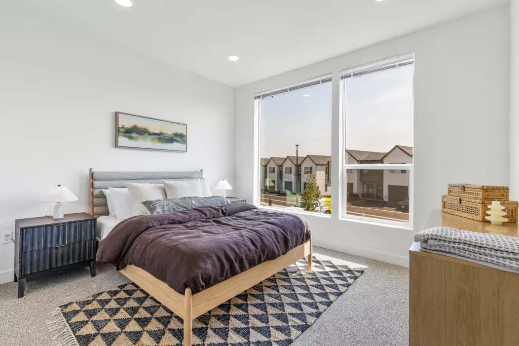 Bright bedroom with a modern wooden bed, plush bedding, and a geometric rug, featuring large windows with a view of neighboring houses.