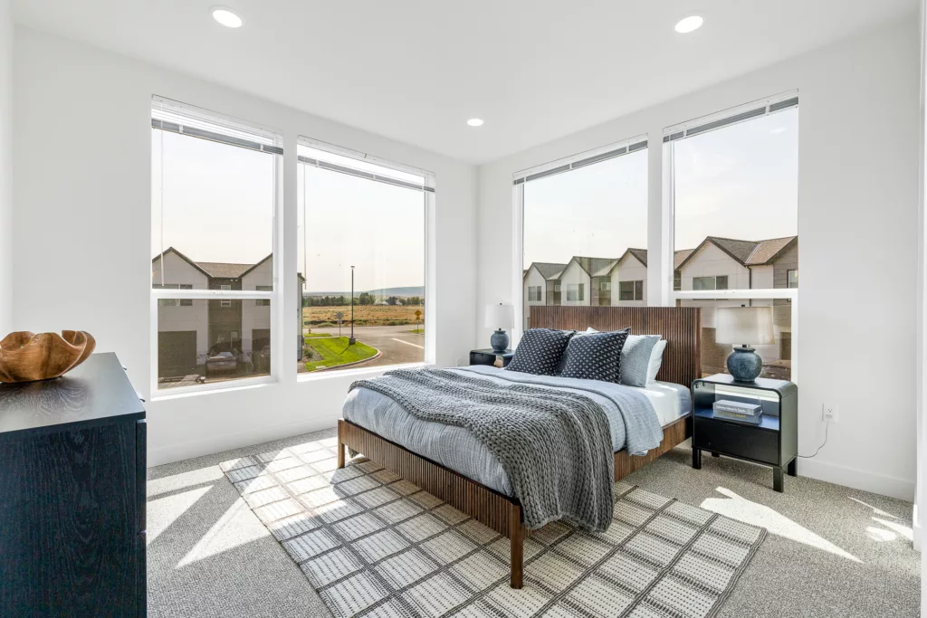 Modern bedroom with large windows, a wooden bed with blue and grey bedding, a dark bedside table with a lamp, and a checkered rug on the floor.