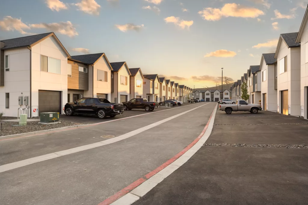A row of modern suburban houses with driveways and parked cars during sunset, viewed from a street lined with neatly paved sidewalks.