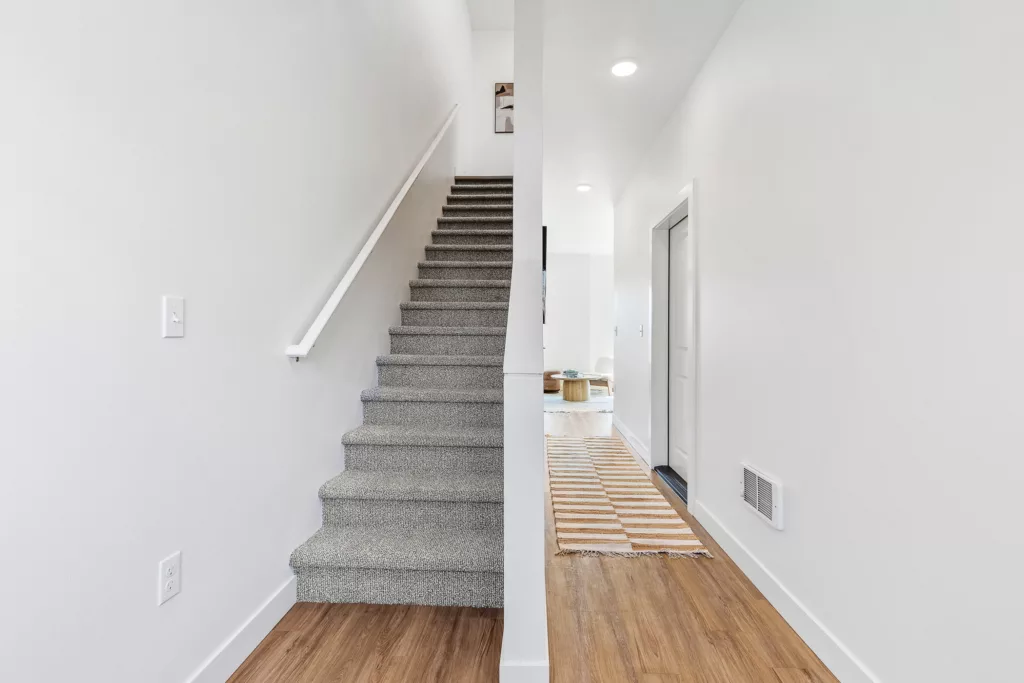 Interior scene showing a carpeted staircase on the left with a white railing and a hallway on the right with wooden flooring and a striped rug, leading to a room with light furnishings.