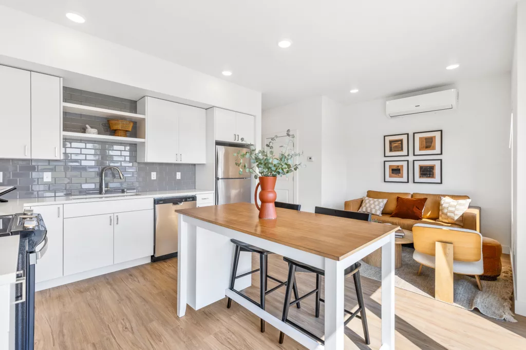 Modern apartment interior featuring a kitchen with white cabinets, gray subway tile backsplash, and stainless steel appliances. A wooden island with high chairs is central, while a cozy living area with a brown sofa and decorative pillows is visible in the background, adorned with framed artwork and a small air conditioning unit.
