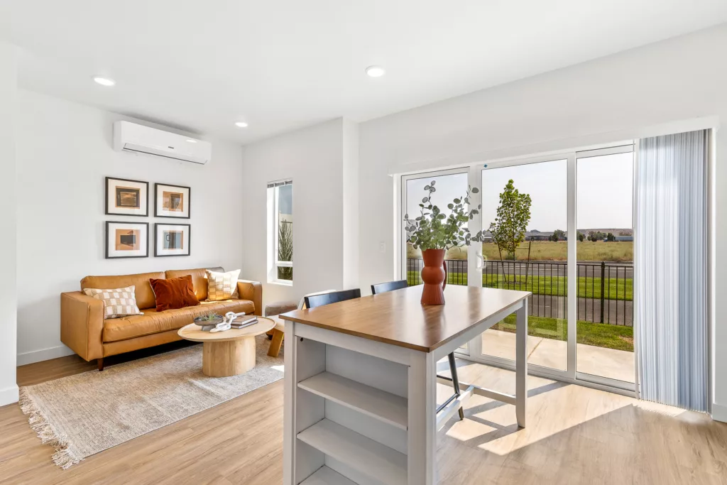 A modern living room with a brown leather sofa, decorative pillows, a rug, and a round wooden coffee table. Adjacent is a dining area featuring a wooden table with a vase of plants, and large sliding glass doors leading to an outdoor view with a lawn and trees.