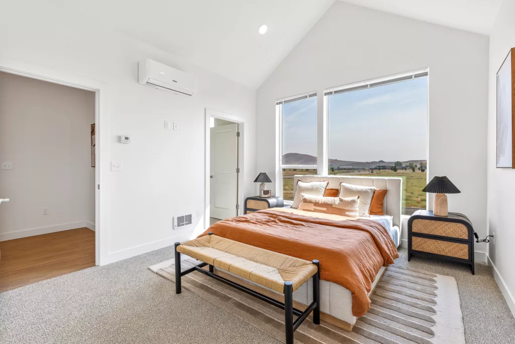 A modern bedroom with a neatly made bed featuring rust-colored bedding and multiple pillows. There are two large windows providing a view of a serene landscape outside. The room includes a small nightstand with a lamp on each side of the bed and a bench at the foot. The walls are white with minimal decor, and the floor is carpeted.