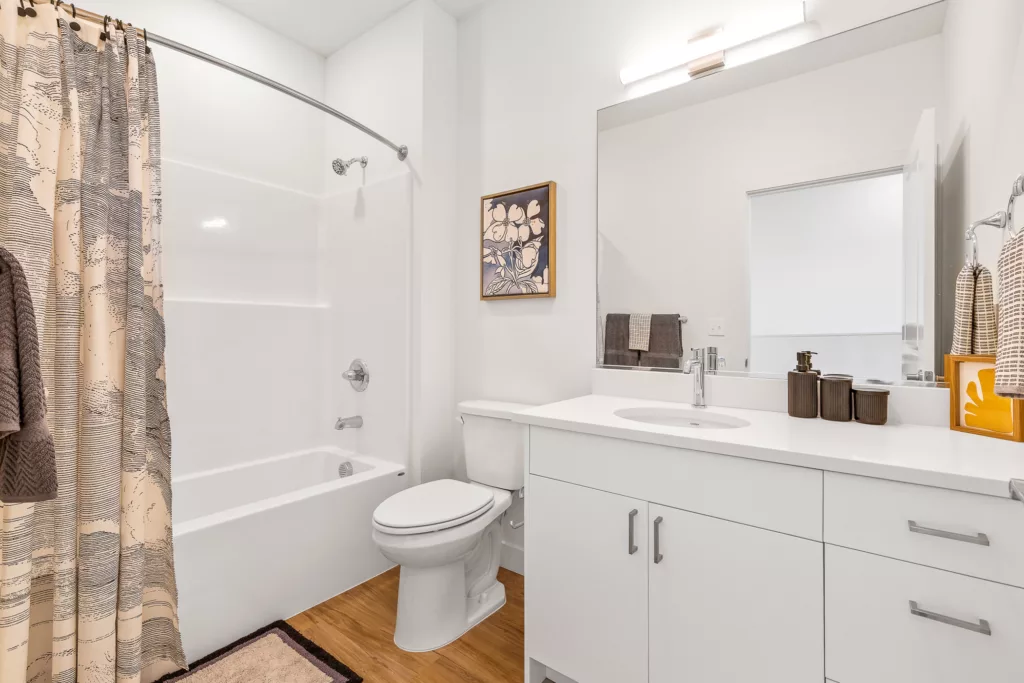 A modern bathroom with a white vanity and round sink, a large wall mirror, wooden floor, and a bathtub with a patterned beige shower curtain. There are dark brown towels, decorative containers on the counter, and a wall-mounted artwork above the toilet.