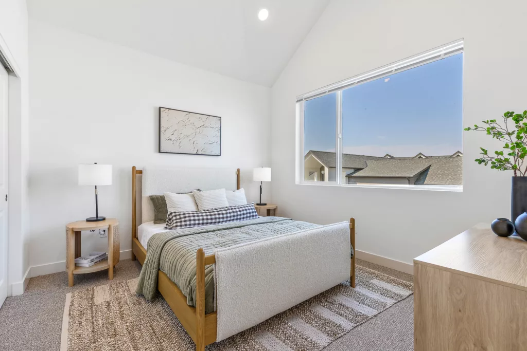 A modern bedroom featuring a bed with white and green bedding, flanked by wooden nightstands with lamps. A large window allows natural light and a view of neighboring rooftops. A textured area rug covers the floor, and a dresser with decorative plants and vases is on the right.