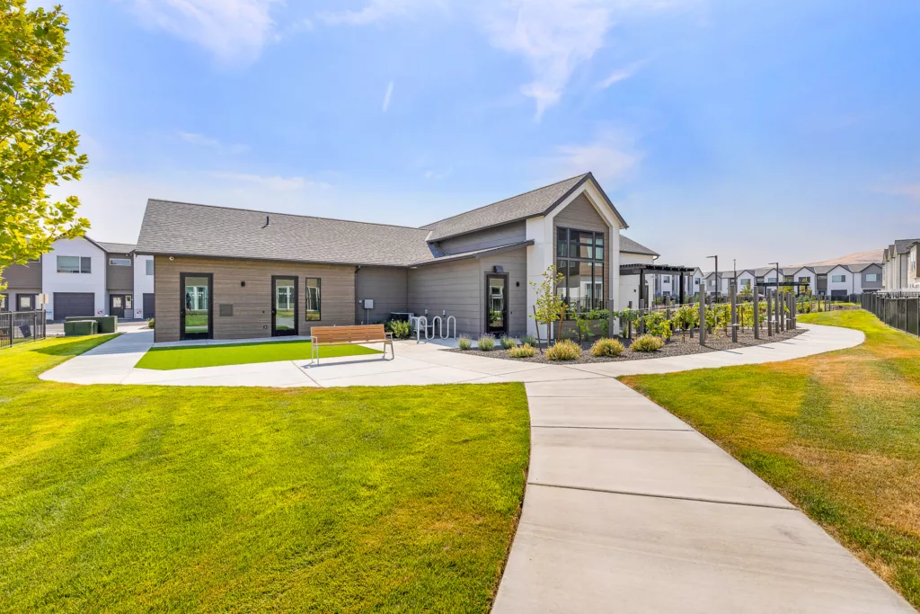 A modern building with a sloped roof, surrounded by a well-maintained lawn and a paved walkway, in a suburban neighborhood. There are bike racks and a bench near the entrance, with smaller houses visible in the background under a clear blue sky.