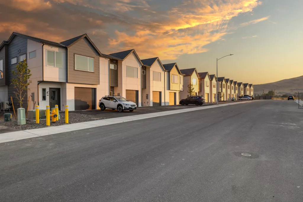 A row of modern two-story houses on a suburban street at sunset, with cars parked in front and dramatic cloud formations in the sky.