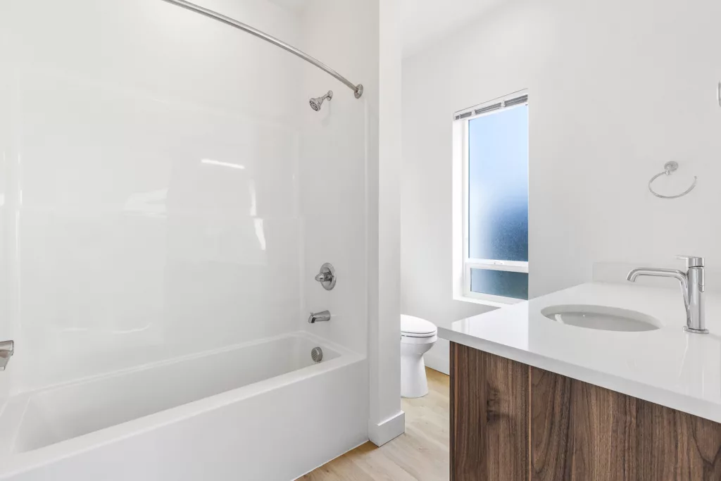 Modern bathroom with a white bathtub and shower, a wooden vanity with a sink, and a frosted window allowing natural light.