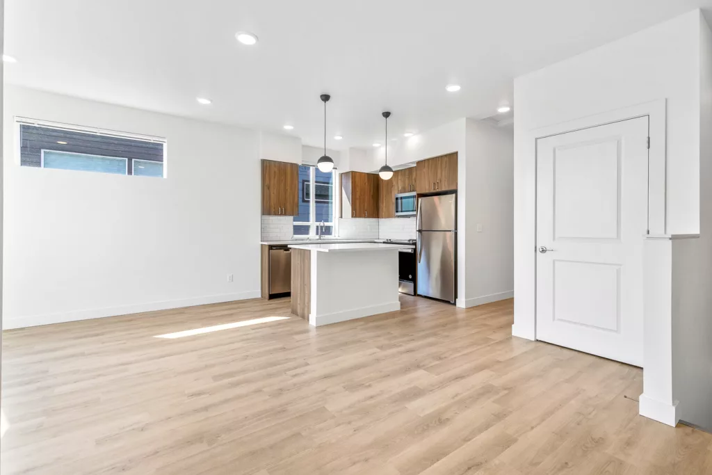 A modern kitchen with wooden floorboards, white walls, and ceiling lights. It features stainless steel appliances, including a refrigerator and dishwasher, wood cabinetry, a white tiled backsplash, and a central island with pendant lighting.