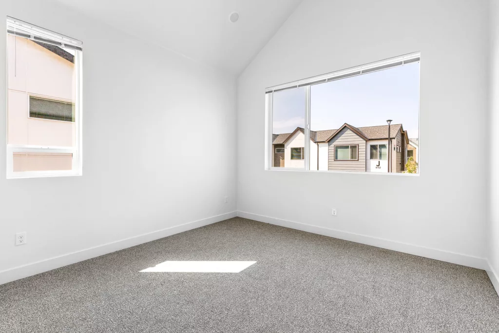 Empty white room with carpeted floor and two windows showing a view of neighboring houses.