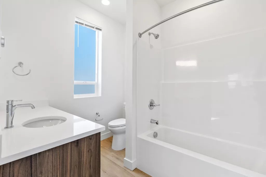 A modern bathroom featuring a white bathtub with a showerhead above, a wooden vanity with a white countertop and chrome faucet, a toilet, and a window with a pull-down blind, all set against white walls and a light wood floor.
