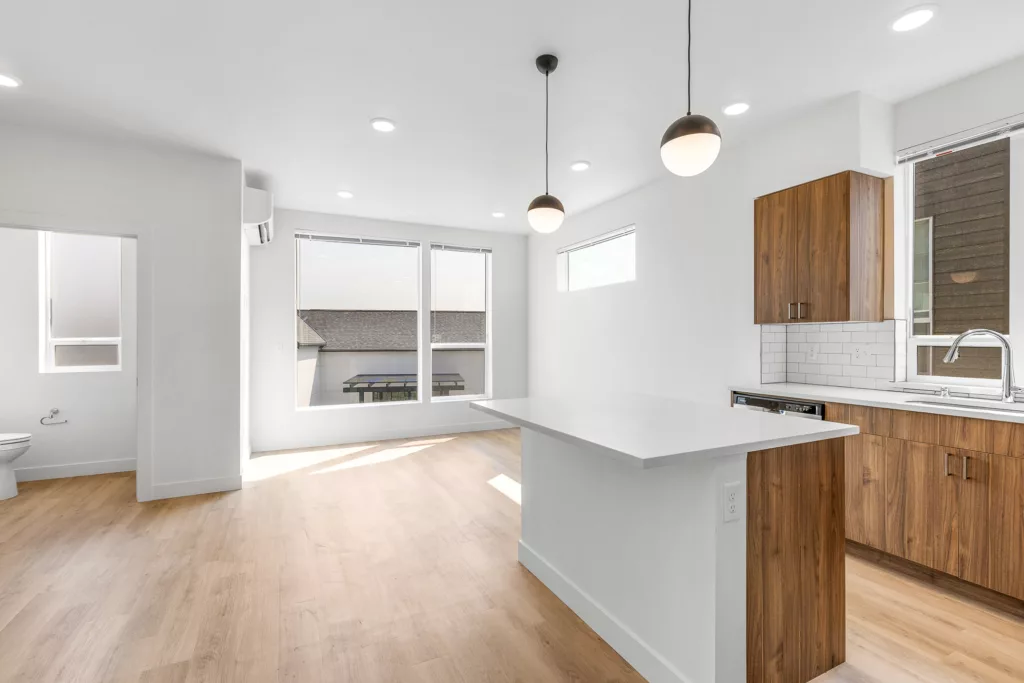 Modern kitchen and dining area with wooden cabinets, a white countertop, and pendant lighting, featuring large windows allowing natural light, and a bathroom visible to the left.