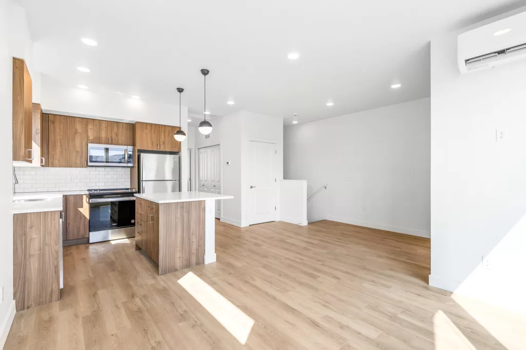 A modern kitchen and living area featuring wooden cabinets, stainless steel appliances, a white island, pendant lights, and light wood flooring.