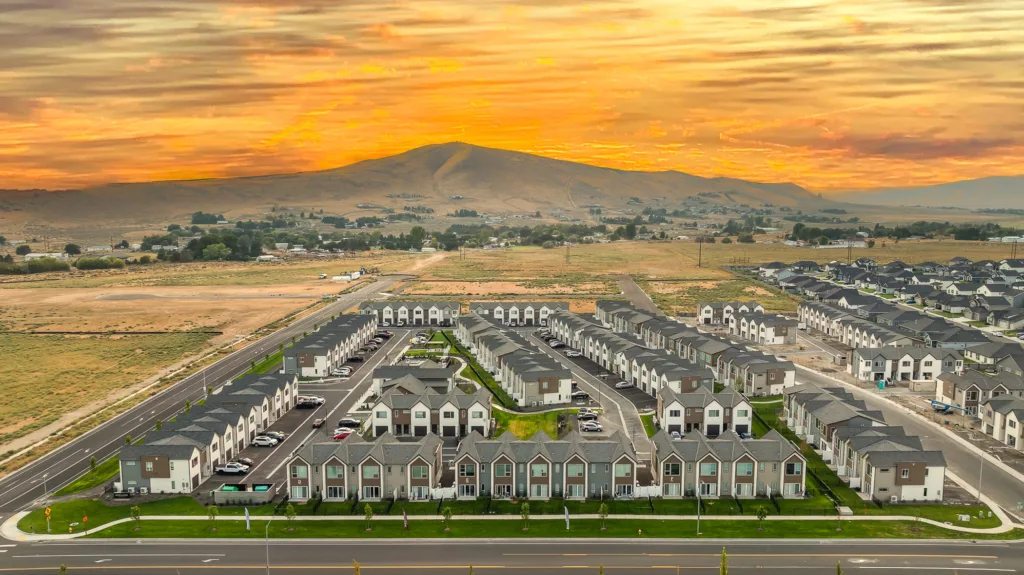 Aerial view of a residential neighborhood comprised of rows of identical two-story houses with gray roofs, surrounded by open fields and roads. A hazy, mountainous landscape is visible in the background under a vibrant orange and yellow sunset sky.