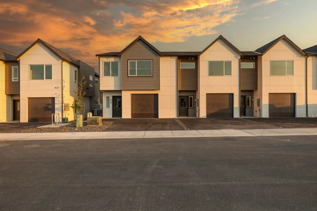 Modern townhouses with identical designs featuring garages, front doors, and large windows, set against a colorful sunset sky.