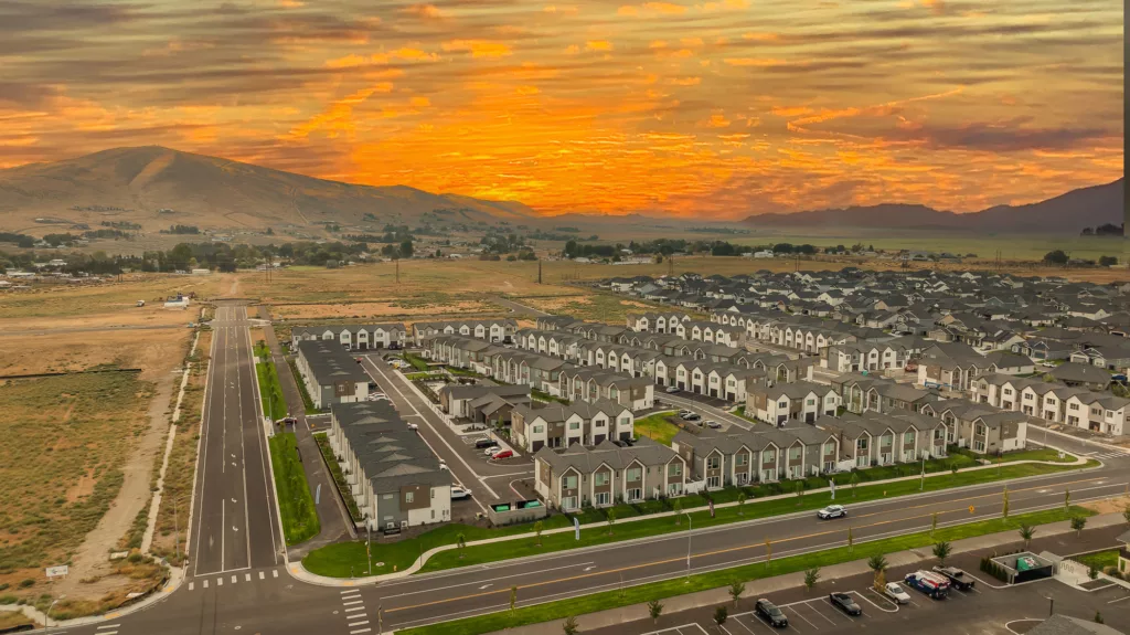 Aerial view of a suburban neighborhood with identical houses, lined streets, and parked vehicles at sunset, with mountains in the background and a vibrant orange sky.