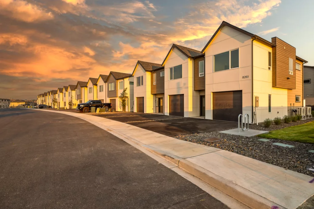 A row of modern two-story townhouses with garages, situated along a wide, empty street at sunset; the sky is painted with orange and pink hues, enhancing the warm tones of the buildings.