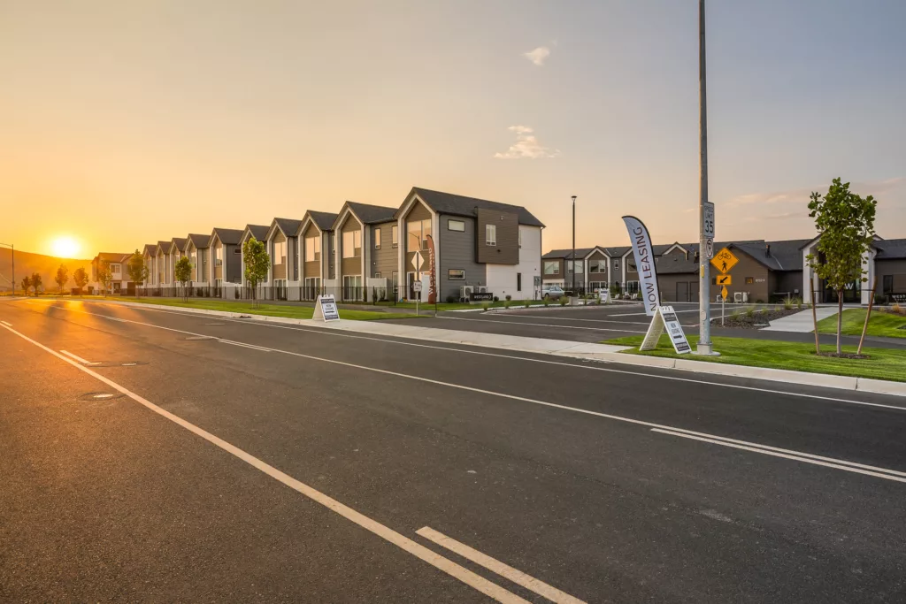 A row of modern townhouses along a street at sunset, with a sign indicating they are now leasing. The sky is golden, and the street is empty, creating a peaceful ambiance.