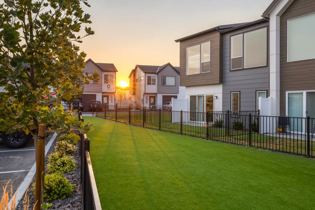 A row of modern two-story houses with large windows, separated by a black metal fence, and a neatly manicured lawn in the foreground, with the sun setting in the background.