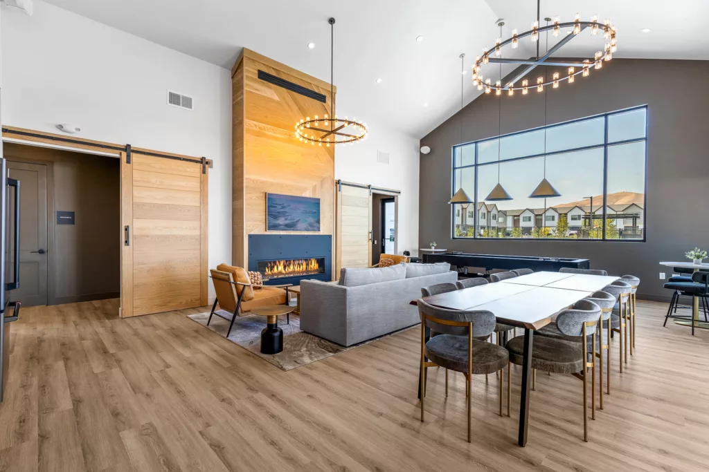 A modern living room featuring a fireplace with a wide mantel, surrounded by a wooden accent wall and sliding barn doors. The room has high ceilings, a large window with a view of neighboring houses, gray sectional couch, two tan leather chairs, and a long dining table with multiple chairs. Two circular chandeliers provide overhead lighting.